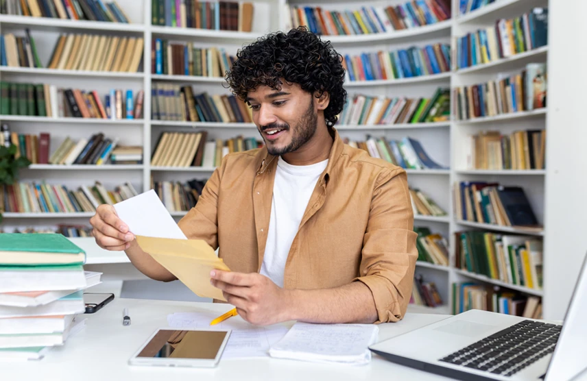 Score Big scholarships, insider tips from EducationQuest. Mail student in library with smile on face as he reviews scholarship award letter.